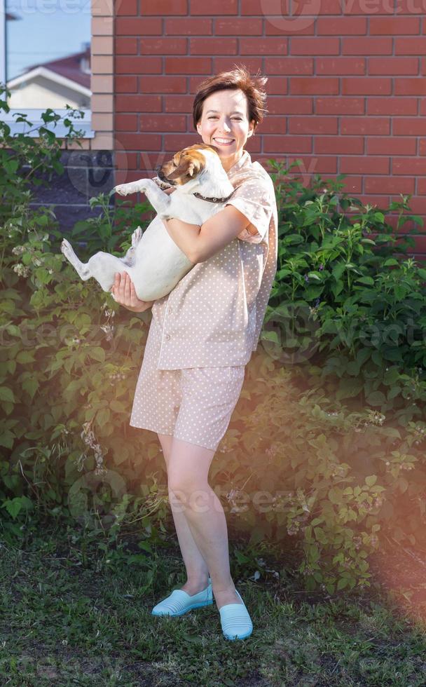 Young woman wearing homewear holding jack russell terrier dog near his house on backyard - pet owner and animal photo
