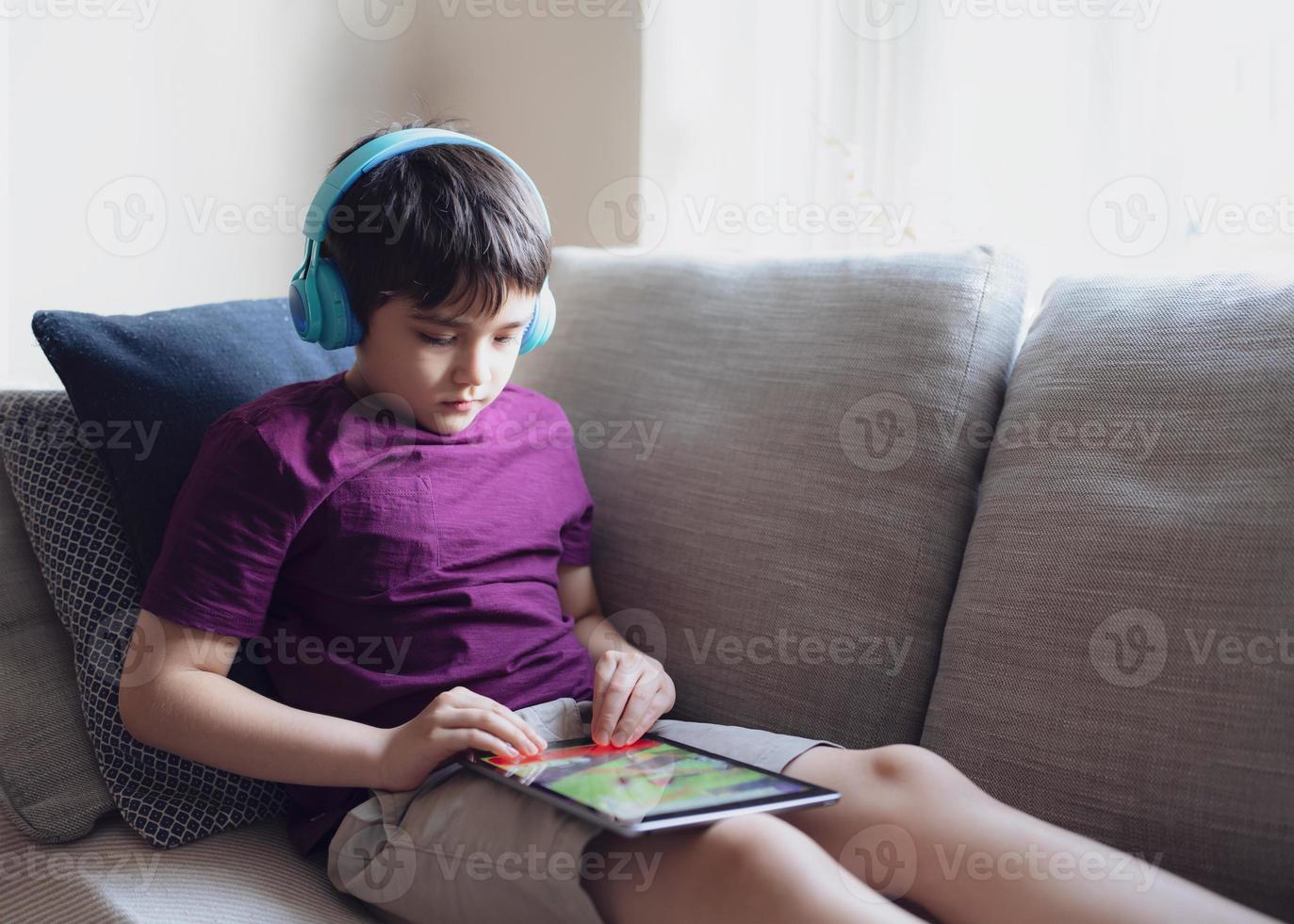 concepto de educación, retrato clave de un niño de escuela primaria leyendo un libro electrónico en una tableta para la tarea, un niño usando auriculares jugando en línea en Internet, un niño sentado en un sofá viendo dibujos animados en una libreta digital foto