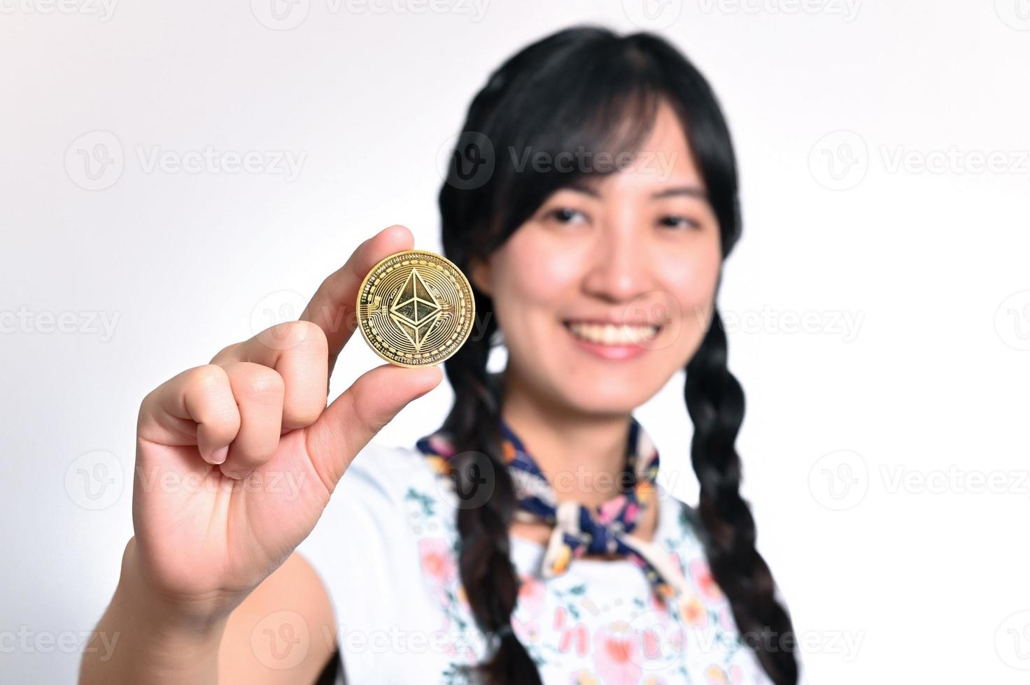 retrato de una hermosa joven asiática feliz vestida de denim sosteniendo una moneda criptográfica sobre fondo blanco. foto