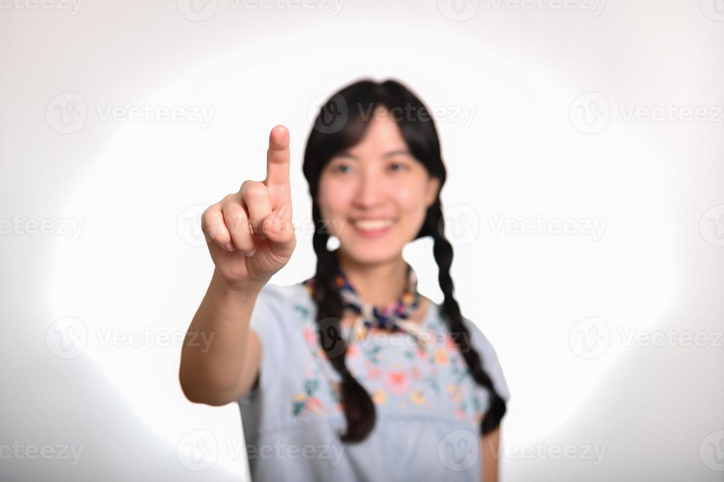 Portrait of beautiful happy young asian woman in denim dress touching invisible screen on white background. photo