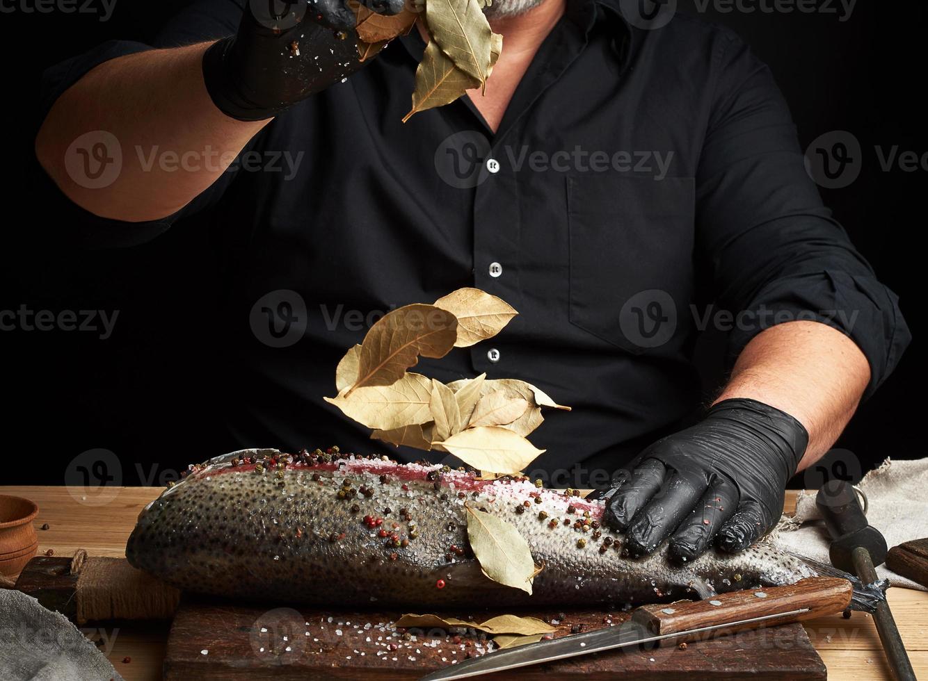 chef con uniforme negro y guantes de látex negros vierte hojas secas de laurel en un filete de salmón fresco cortado foto