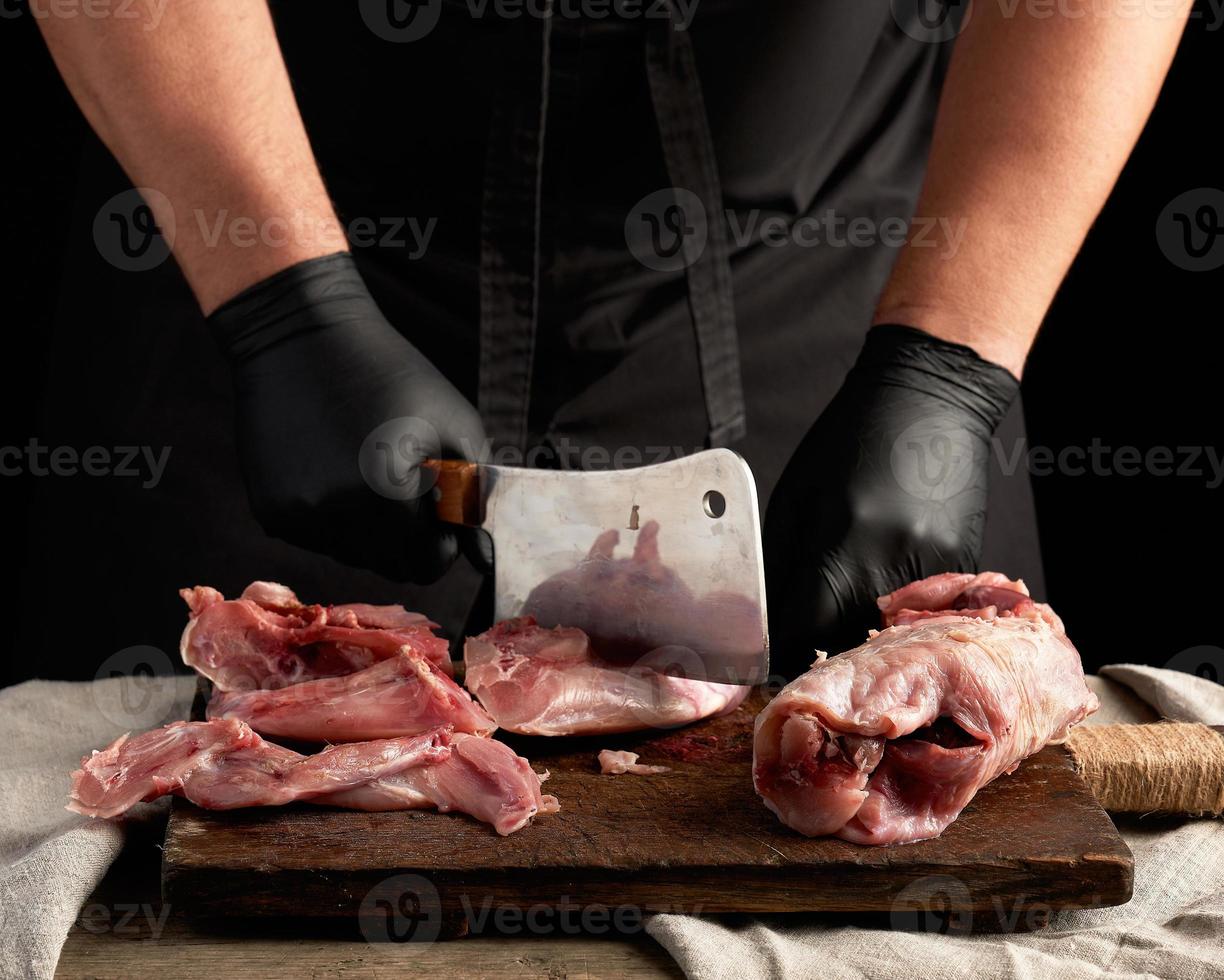 chef in black latex gloves holds a big knife and cuts into pieces raw rabbit meat photo