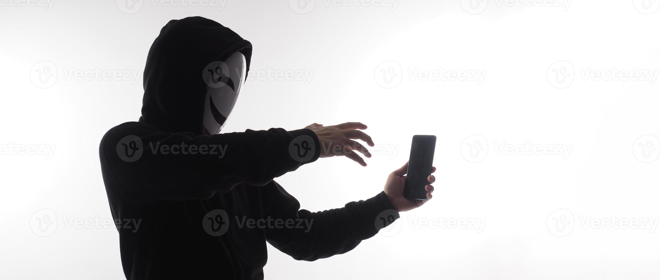 Hacker Anonymous and face mask with smartphone in hand. Man in black hood shirt holding and using mobile phone on white background. Represent cyber crime data hacking or stealing personal data concept photo