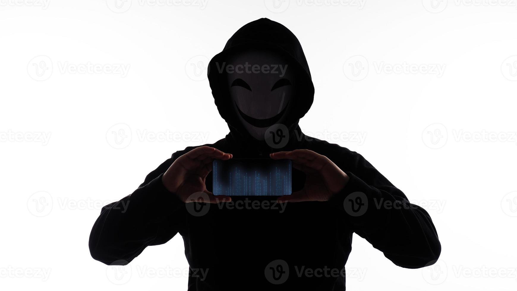 hacker anónimo y mascarilla con smartphone en la mano. hombre con camisa de capucha negra sosteniendo y usando un teléfono móvil con fondo blanco. representan el concepto de piratería informática o robo de datos personales foto