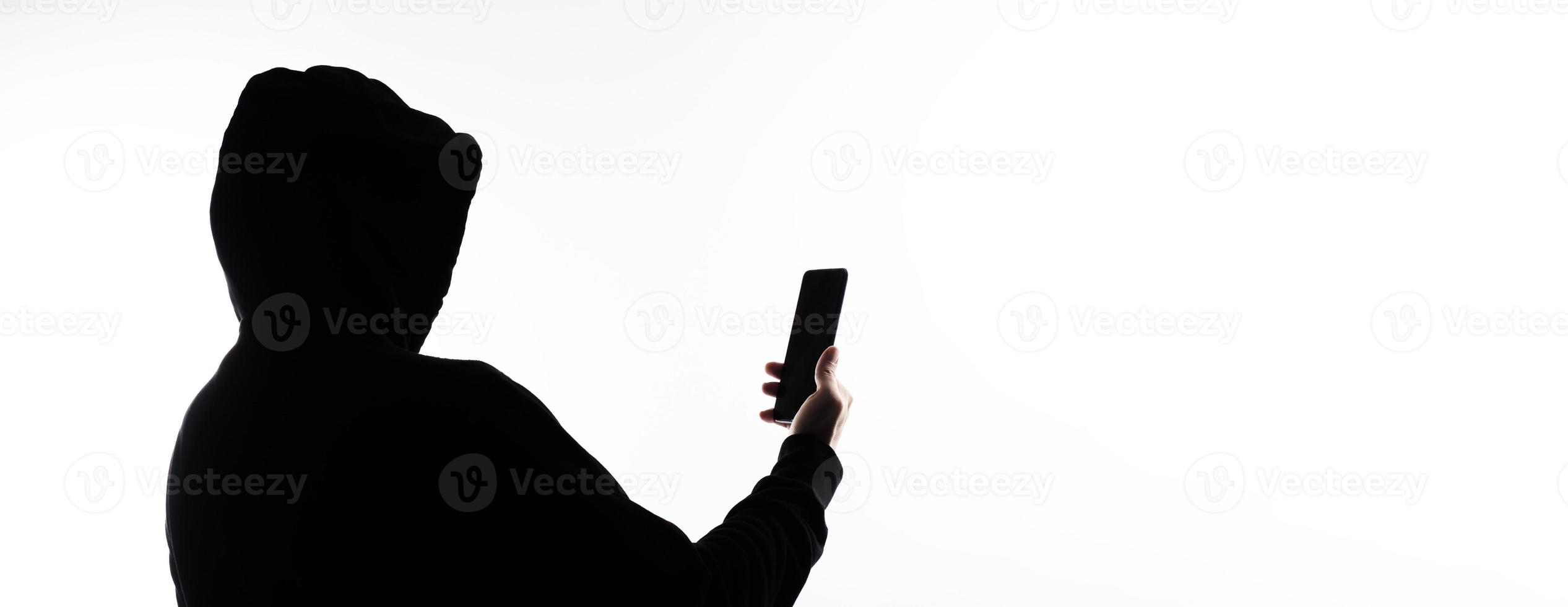 Hacker Anonymous and face mask with smartphone in hand. Man in black hood shirt holding and using mobile phone on white background. Represent cyber crime data hacking or stealing personal data concept photo