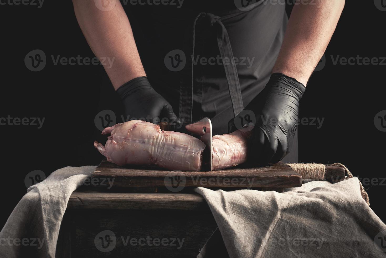 chef in black latex chunks cuts into pieces raw rabbit meat on a brown wooden board photo