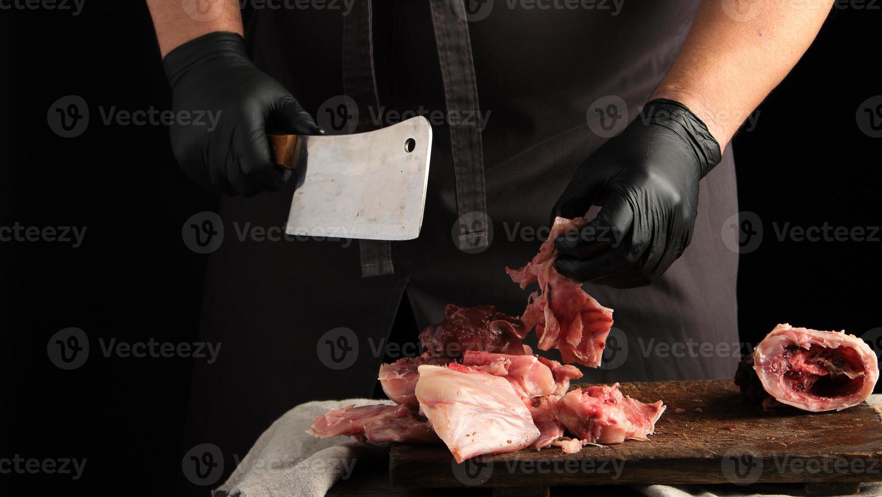 chef in black latex gloves holds a big knife and cuts into pieces raw rabbit meat on a brown wooden cutting board photo