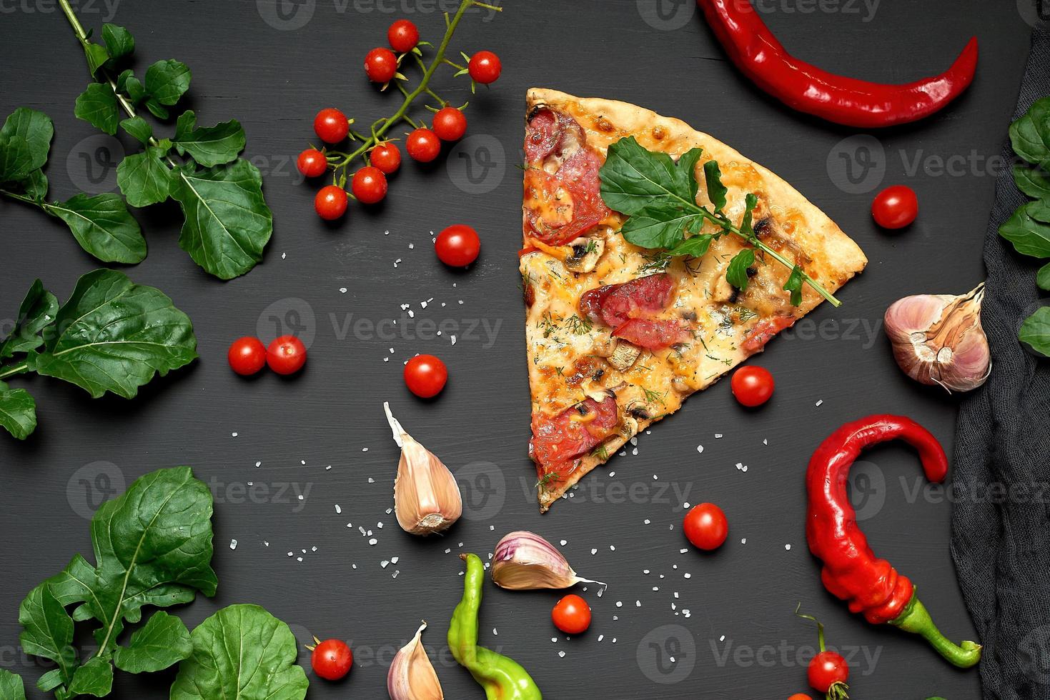triangular slice of baked pizza with mushrooms, smoked sausages, tomatoes and cheese, next to fresh green leaves of arugula photo