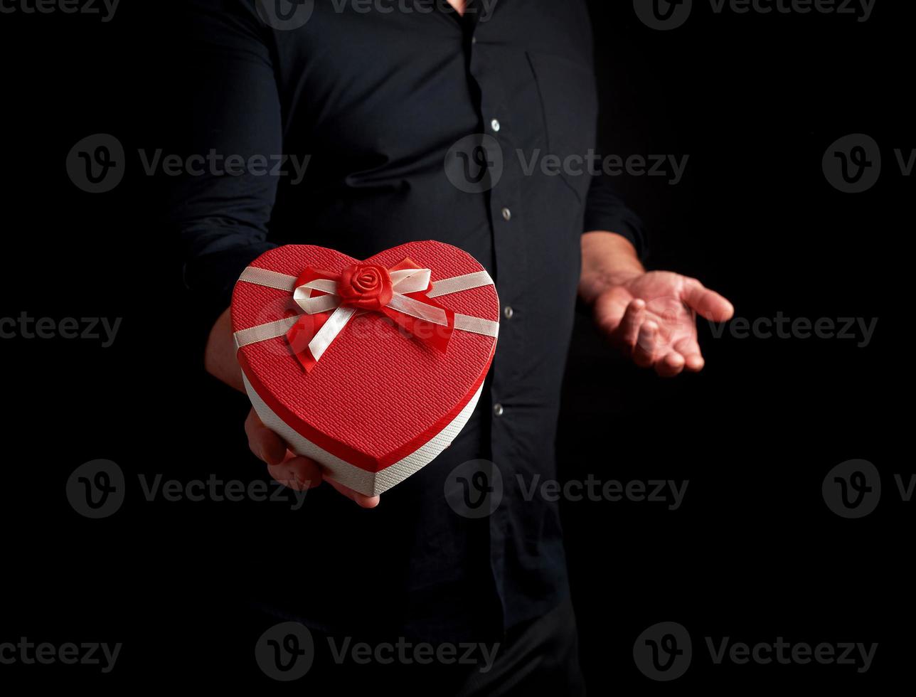 adult man in a black shirt holds a red cardboard box in the form of a heart with a bow on a dark background photo