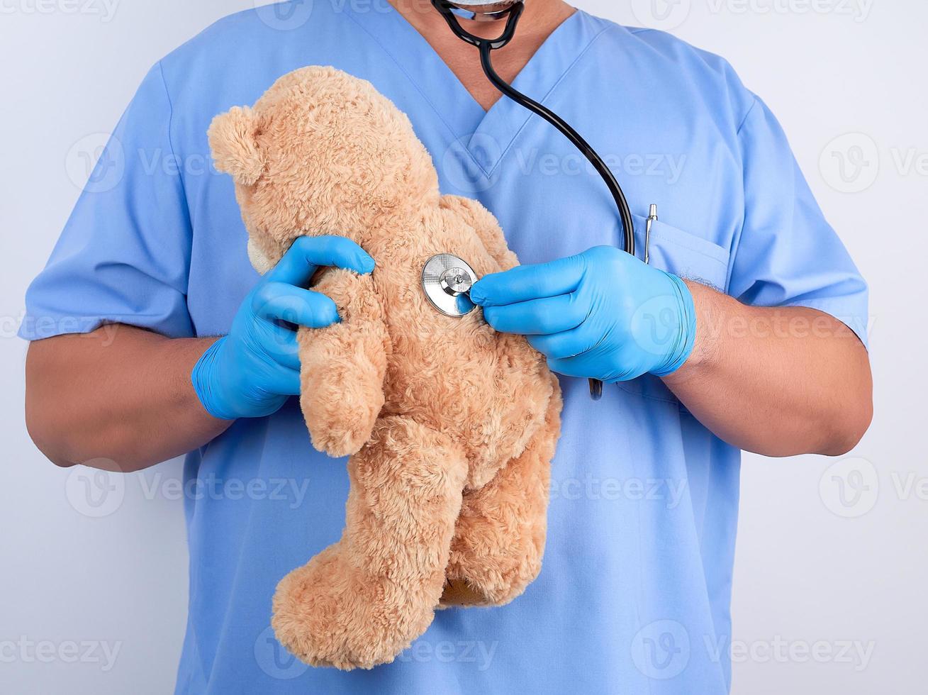 médico con uniforme azul y guantes de látex blancos sosteniendo un oso de peluche marrón foto