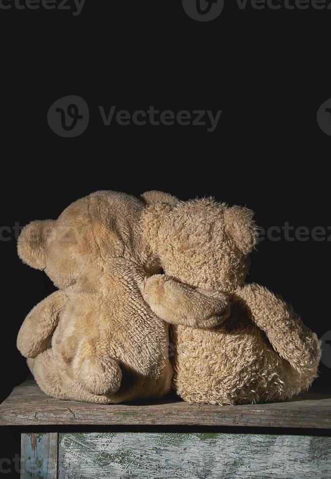 two old brown teddy bears sit hugging on a wooden surface photo