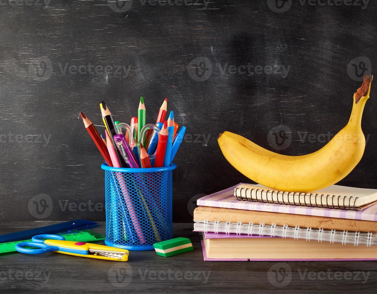 yellow banana on a stack of notebooks, a blue stationery glass with multi-colored wooden pencils photo