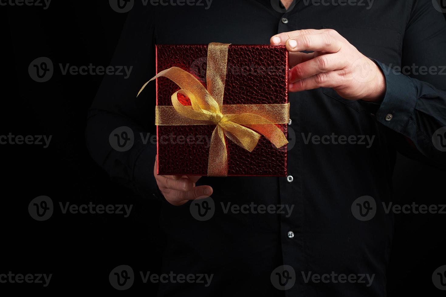 adult man in a black shirt holds a red square box with a knotted golden bow on a dark background photo