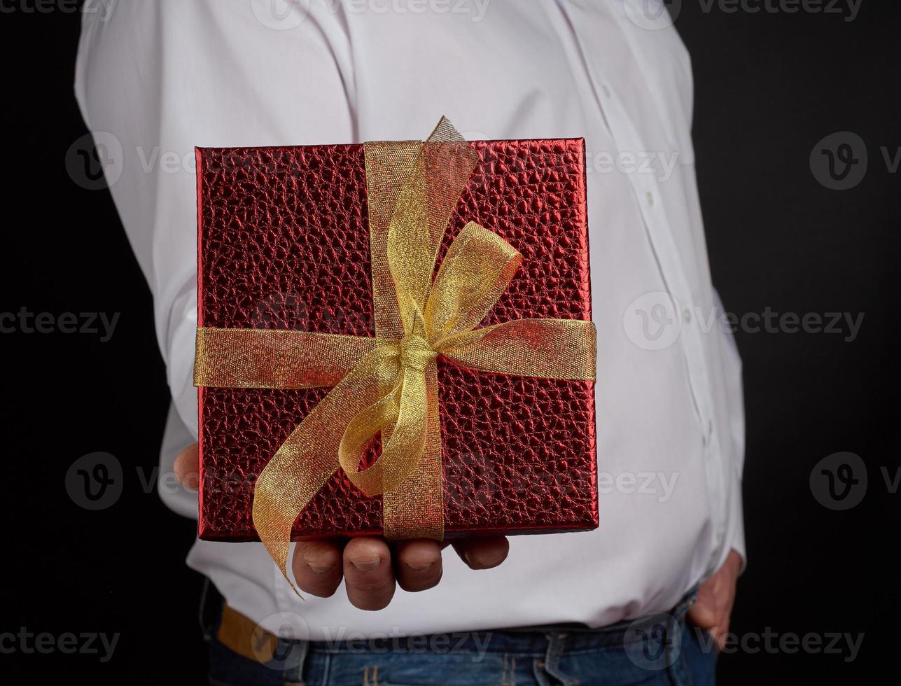 adult man in a white shirt holds a red square box with a knotted golden bow on a dark background photo