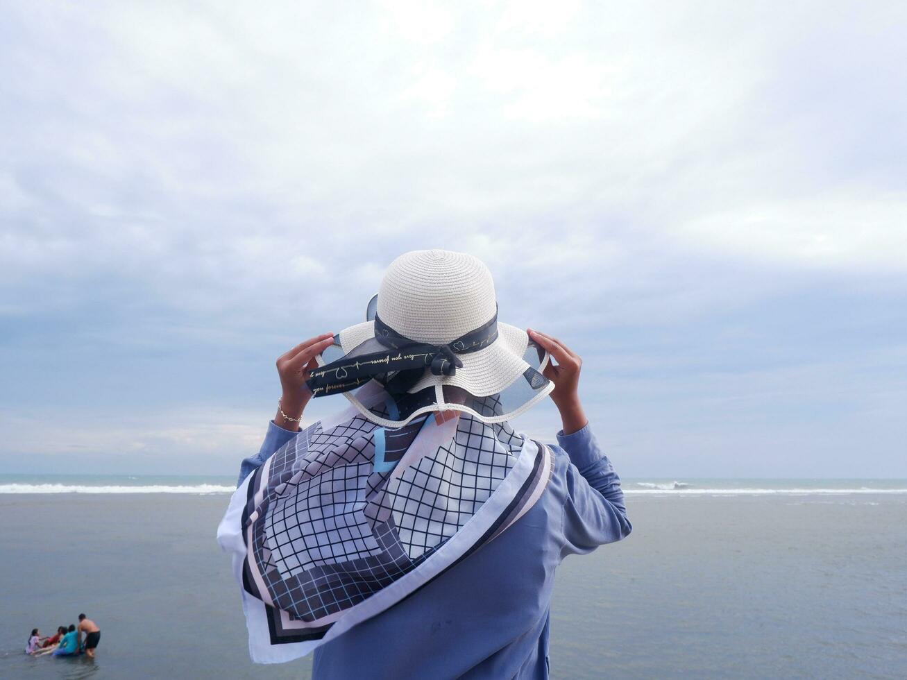 espalda de la mujer en el sombrero en la playa tropical que está mirando el cielo y el mar mientras sostiene su sombrero foto