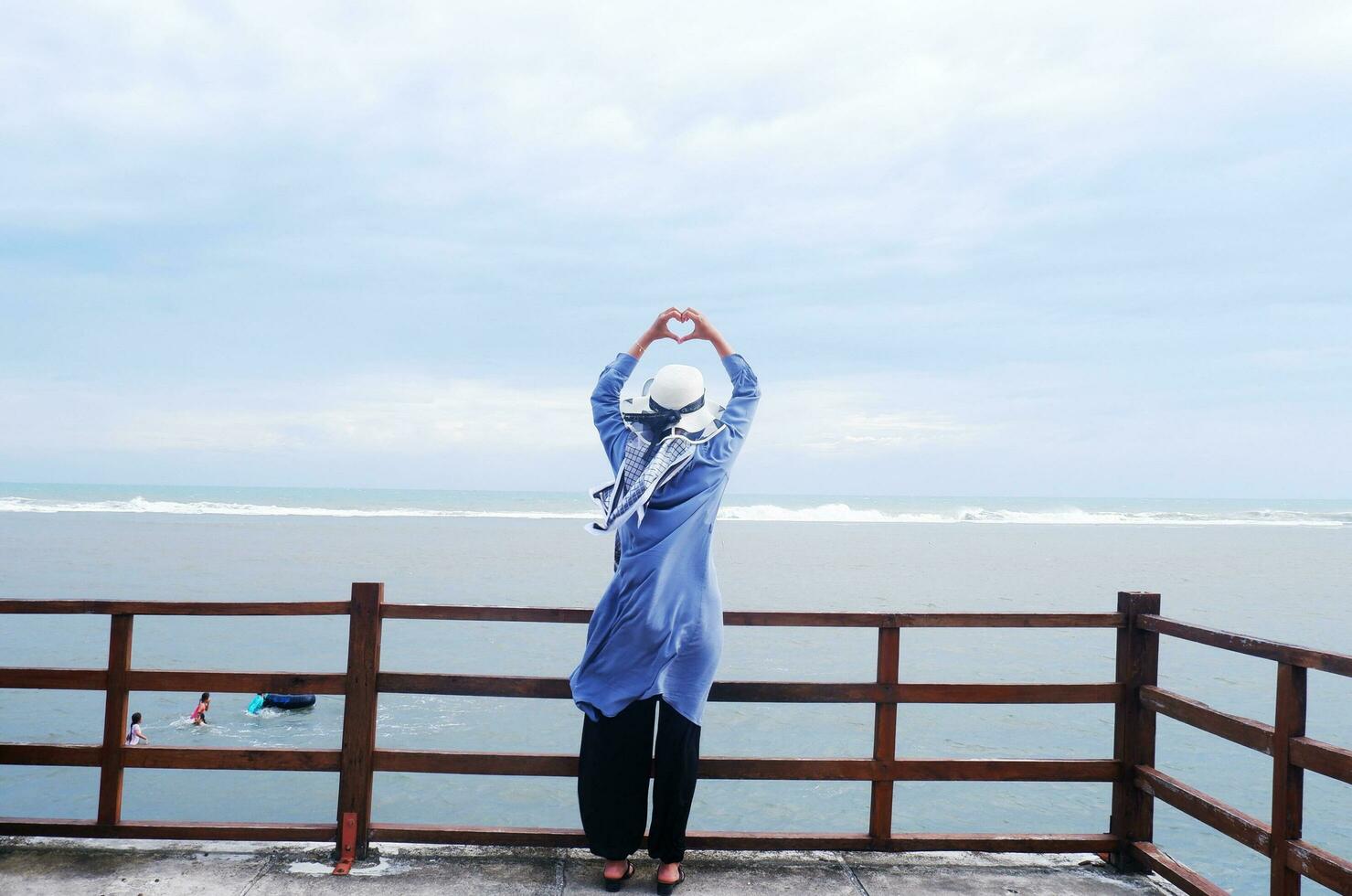 Back of the woman in the hat on the tropical beach who was looking up at the sky and the sea while her hands formed love from the bridge. Beach view photo