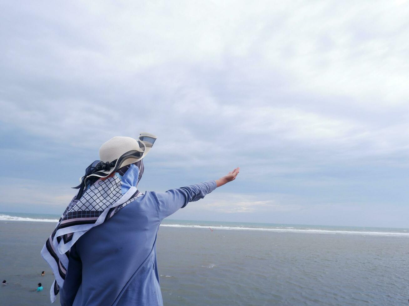 espalda de la mujer en el sombrero en la playa tropical que está mirando el cielo y el mar mientras levanta la mano en oración foto