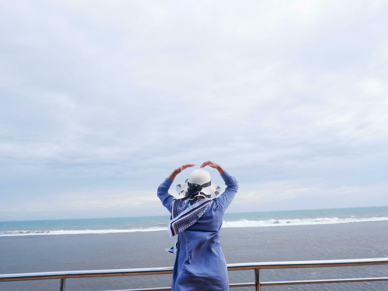 Back of the woman in the hat on the tropical beach who was looking up at the sky and the sea while her hands formed love from the bridge. Beach view photo