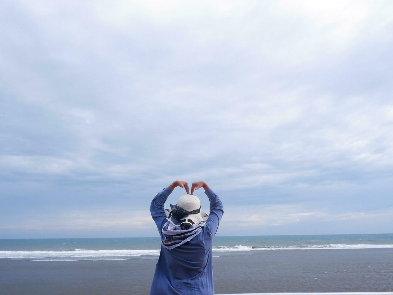 Back of the woman in the hat on the tropical beach who is looking up at the sky and the sea while her hands form love photo