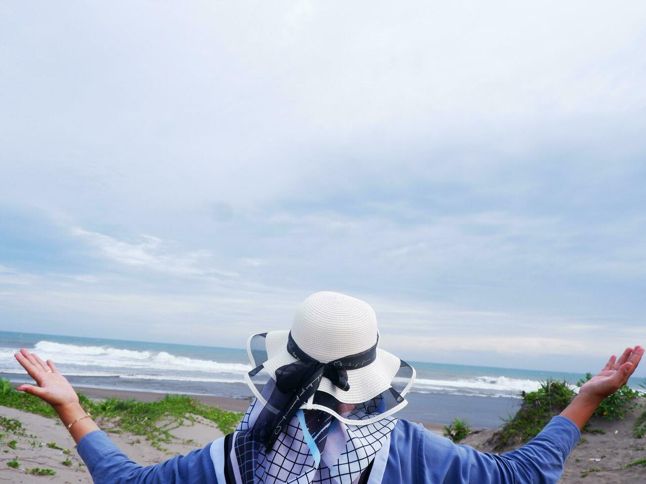 la parte de atrás de la mujer con sombrero en la playa tropical que mira el cielo y el mar mientras extiende sus manos foto