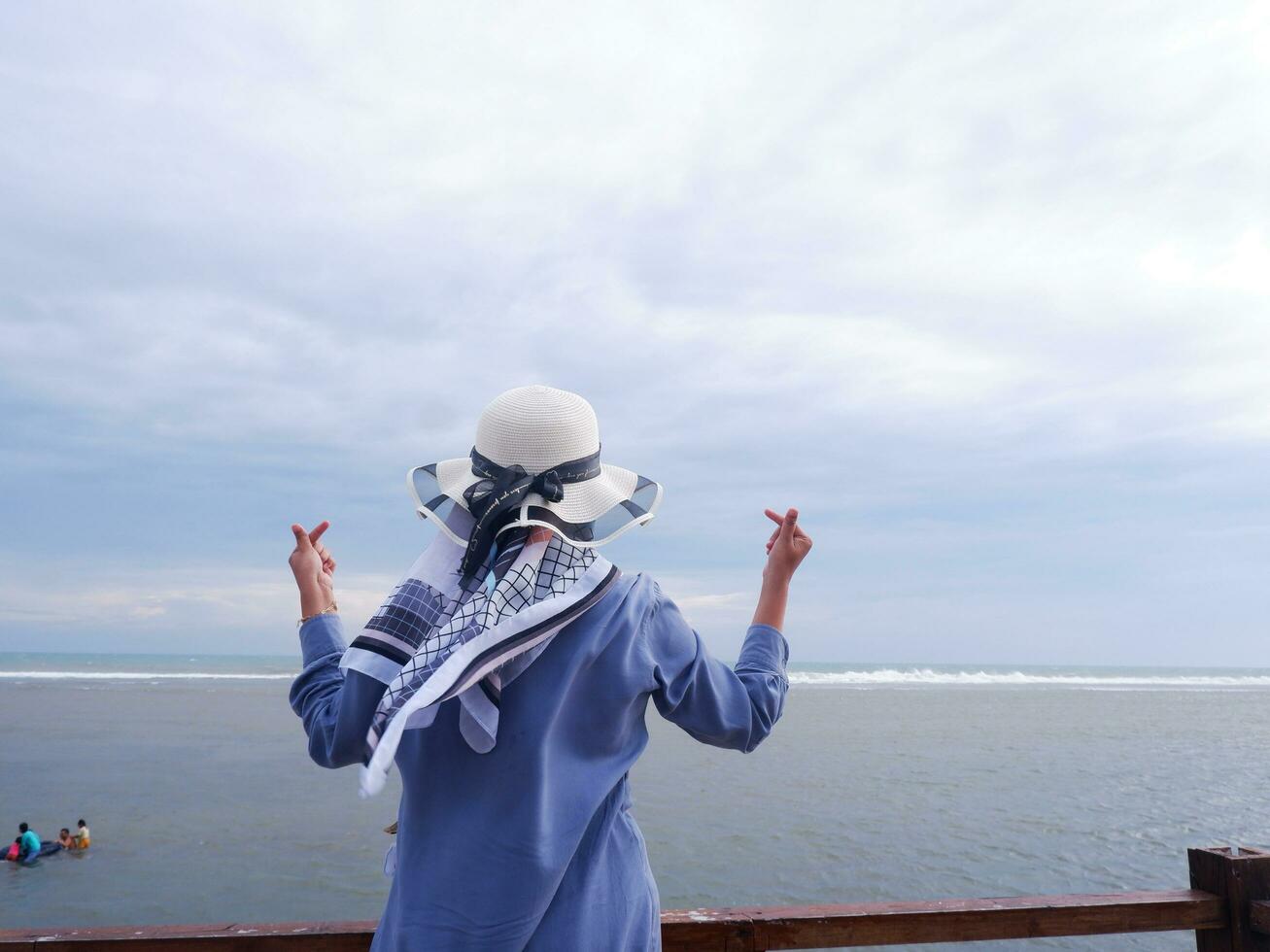 Back of the woman in the hat on the tropical beach who was looking up at the sky and the sea while her hands formed love from the bridge. Beach view photo