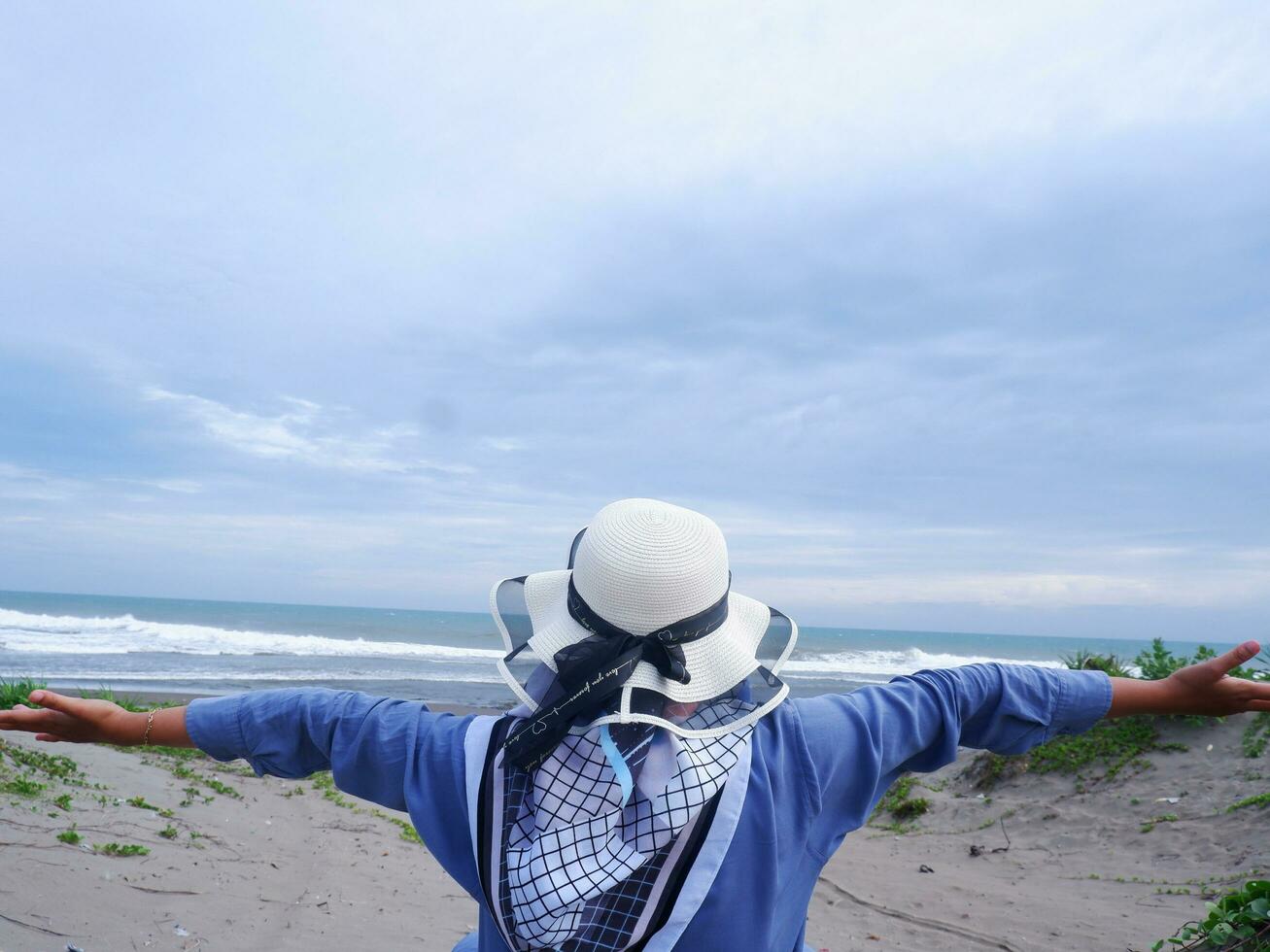 The back of the woman in the hat on the tropical beach who is looking at the sky and the sea while spreading her hands photo