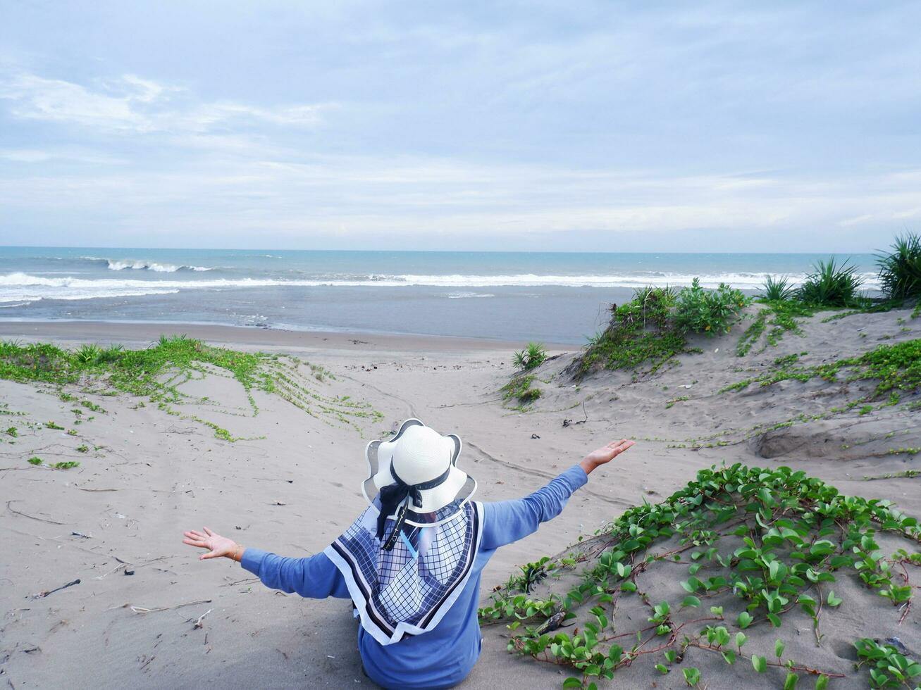 Back of woman in the hat on the tropical beach who was sitting on the sand and looking at the sky and the sea, while spreading her arms . happy women.Beach view photo