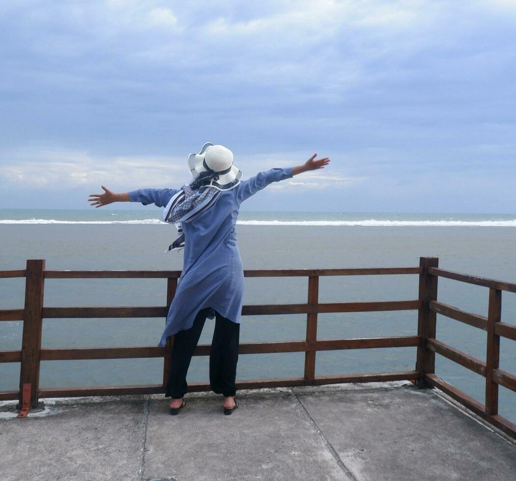 Back of the woman in the hat on the tropical beach who was looking at the sky and the sea while spreading her hands on the bridge. Sea view photo