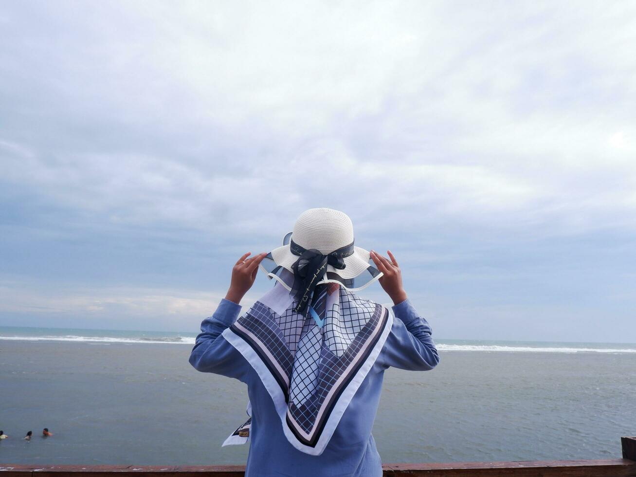 Back of the woman in the hat on the tropical beach who is looking at the sky and the sea while holding her hat photo