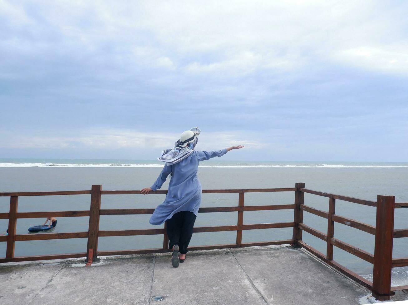 Back of the woman in the hat on the tropical beach who was looking at the sky and the sea while spreading her hands on the bridge. Sea view photo