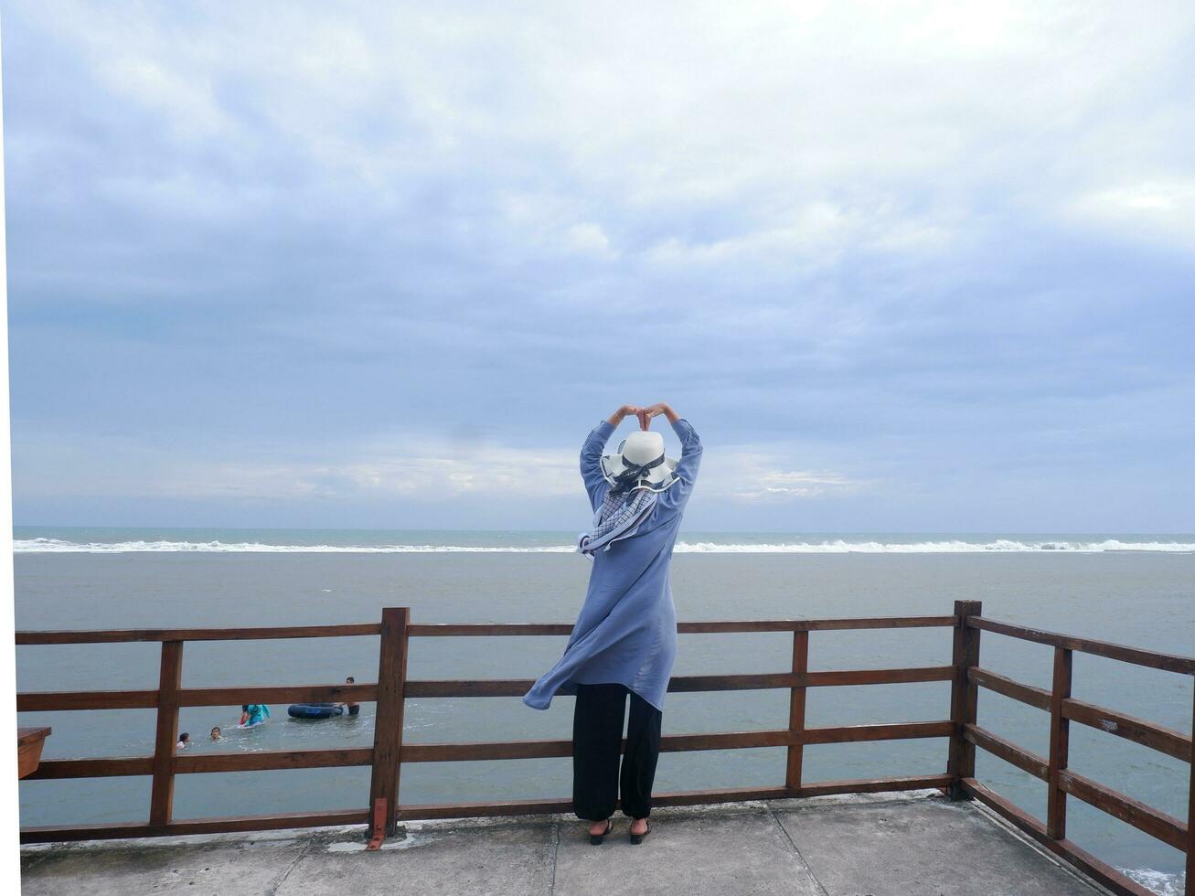 Back of the woman in the hat on the tropical beach who was looking up at the sky and the sea while her hands formed love from the bridge. Beach view photo