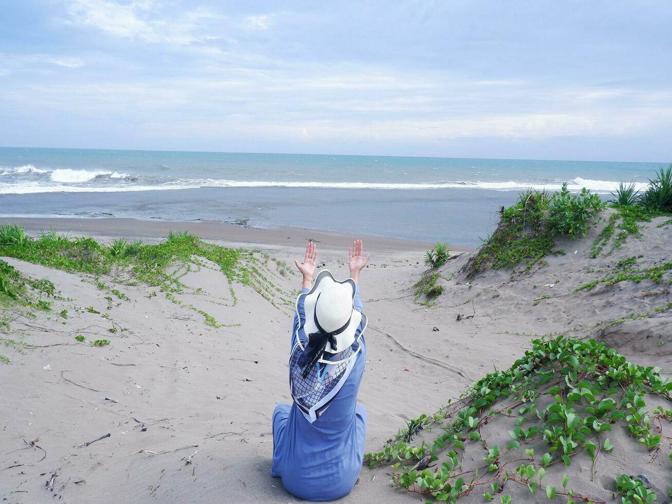 espalda de mujer con sombrero en la playa tropical que estaba sentada en la arena y mirando al cielo y al mar, mientras extendía los brazos. mujer feliz vista a la playa foto