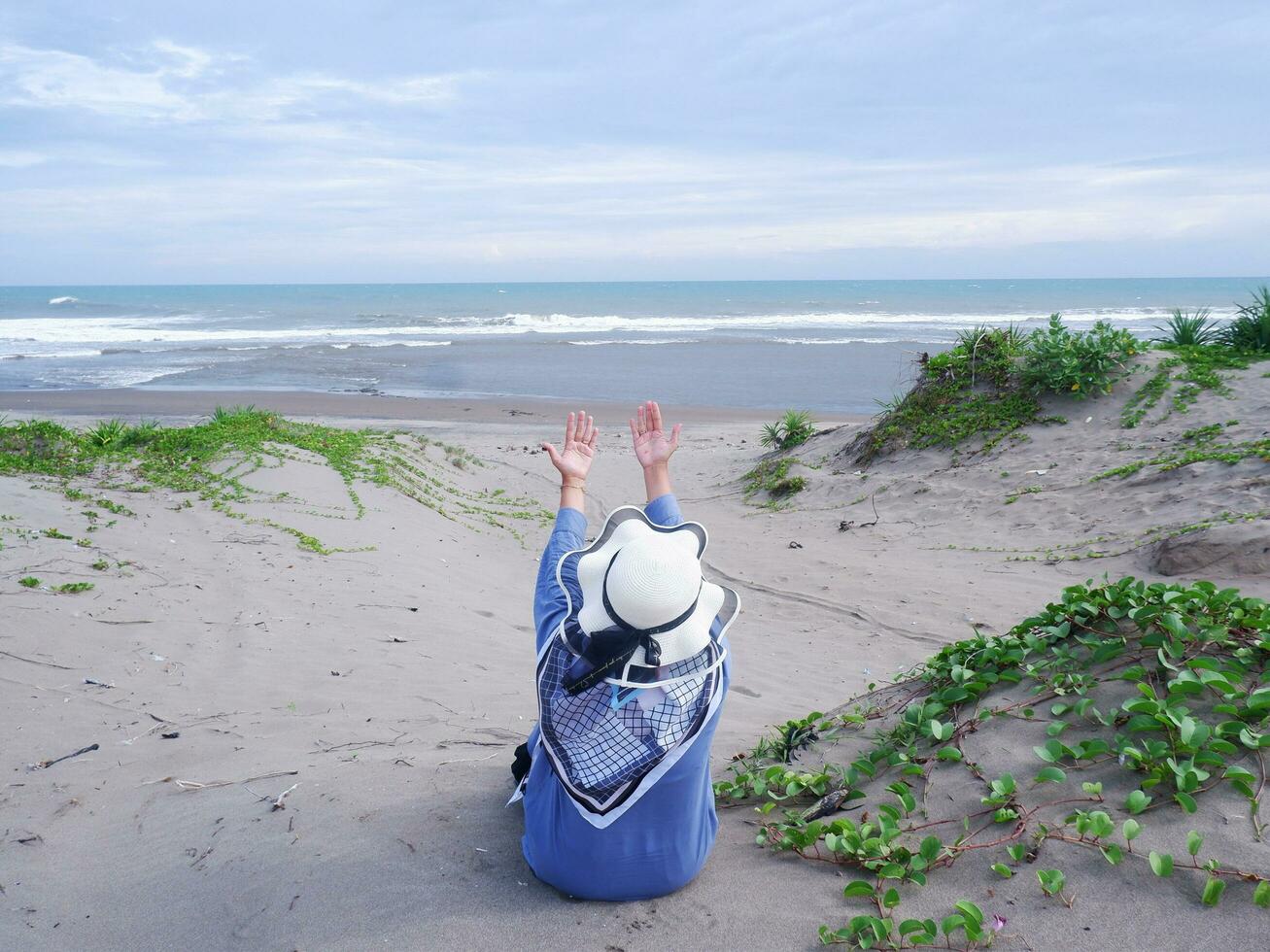 Back of woman in the hat on the tropical beach who was sitting on the sand and looking at the sky and the sea, while spreading her arms . happy women.Beach view photo