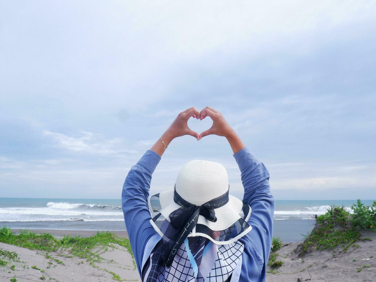 Back of the woman in the hat on the tropical beach who is looking up at the sky and the sea while her hands form love photo