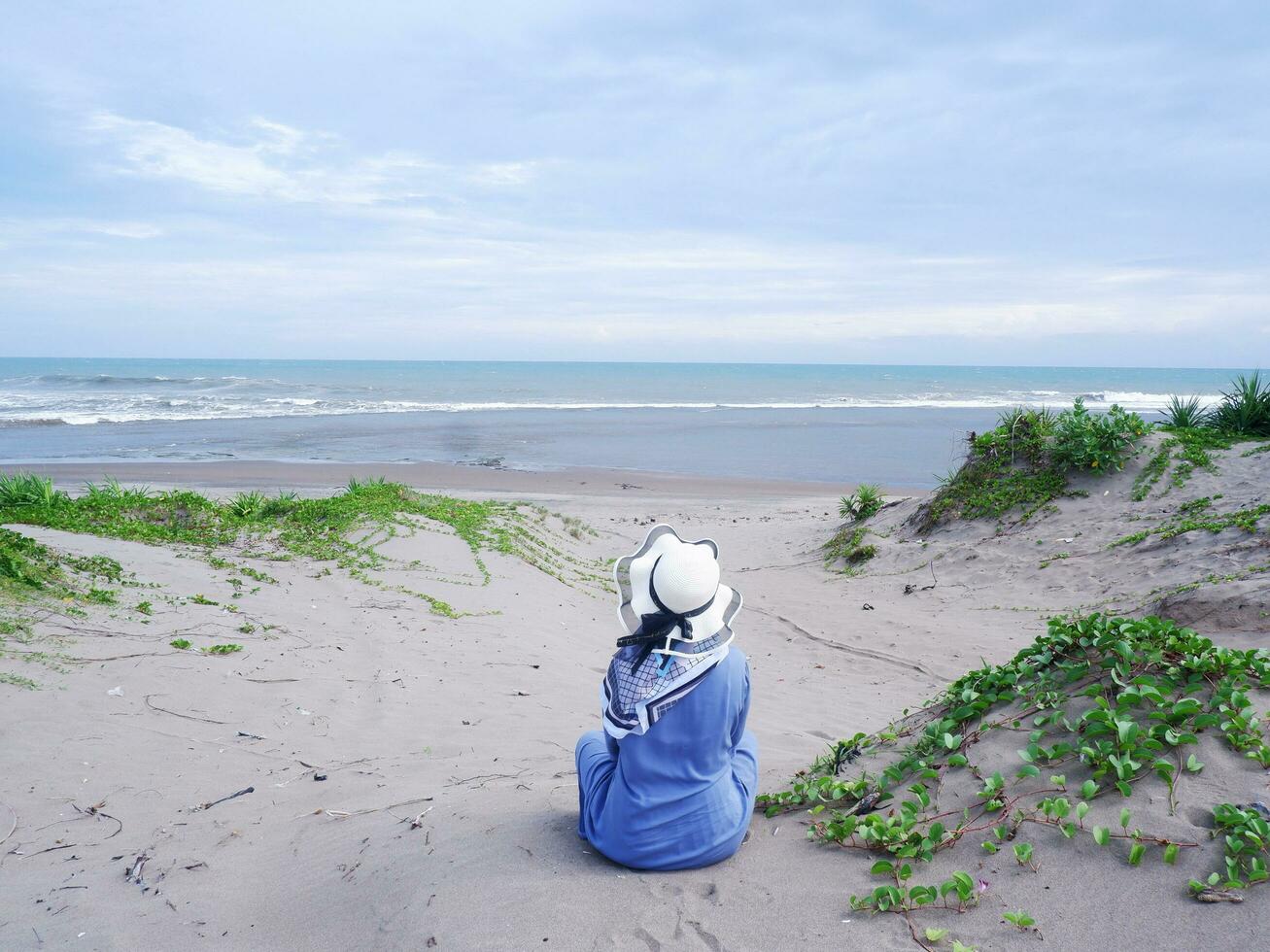 Back of the woman in the hat on the tropical beach who was sitting on the beach sand. background of beach sand and sky. sand mountain.Beach View photo