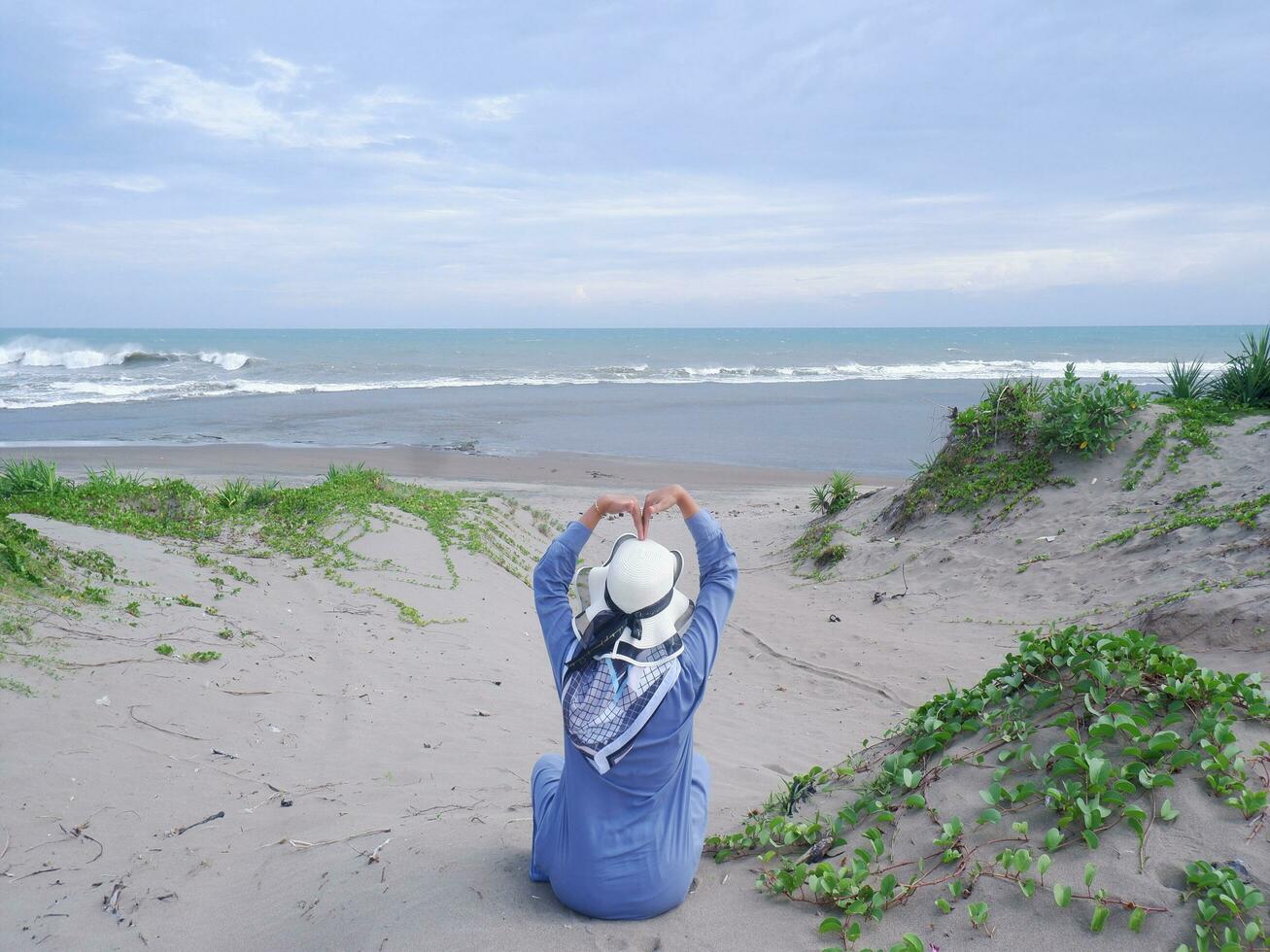 la espalda de la mujer con sombrero en la playa tropical que estaba sentada en la arena de la playa y mirando el océano y luego formó el amor de la mano. fondo de arena de playa y cielo. montaña de arena foto