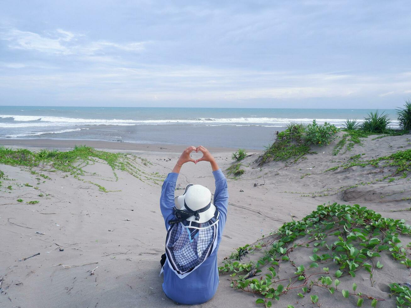 la espalda de la mujer con sombrero en la playa tropical que estaba sentada en la arena de la playa y mirando el océano y luego formó el amor de la mano. fondo de arena de playa y cielo. montaña de arena foto