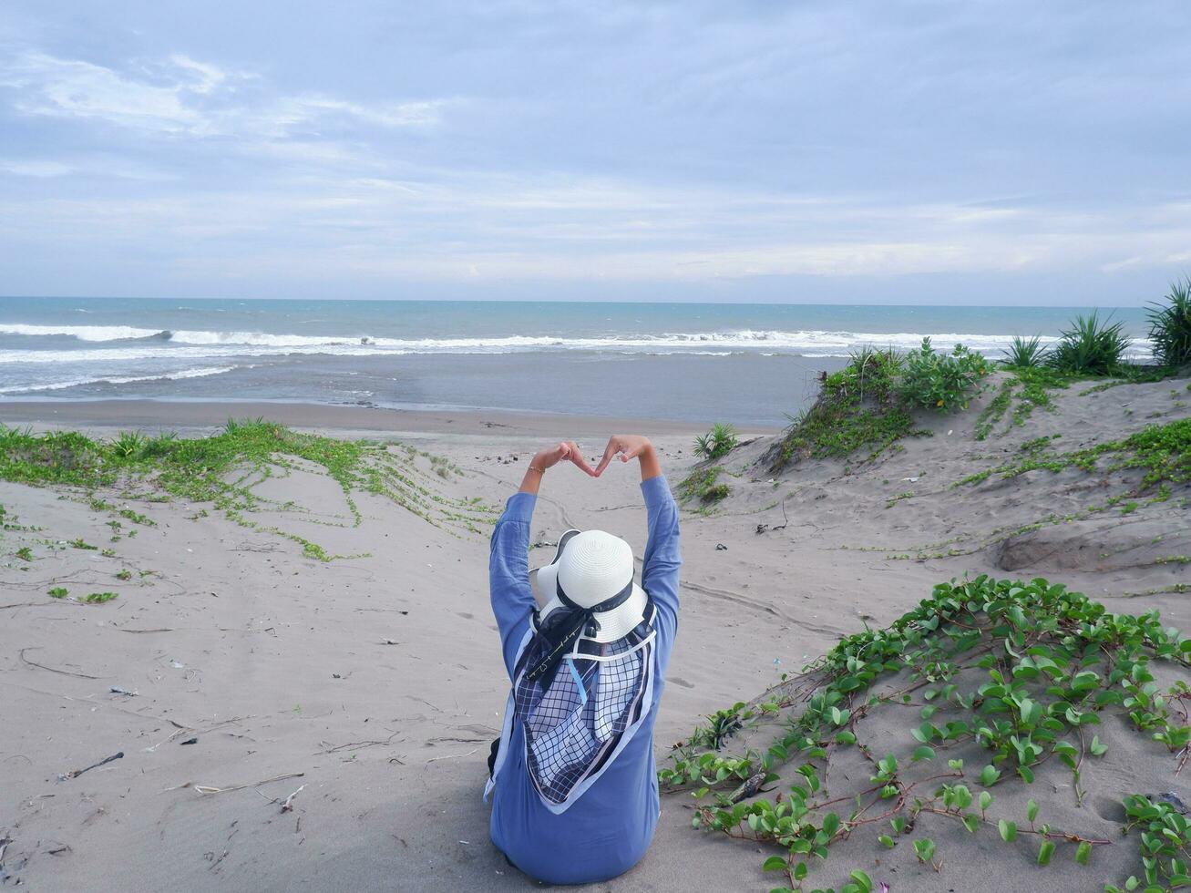 Back of the woman in the hat on the tropical beach who was sitting on the beach sand and staring at the ocean then formed love from the hand. background of beach sand and sky. Sand Mountain photo