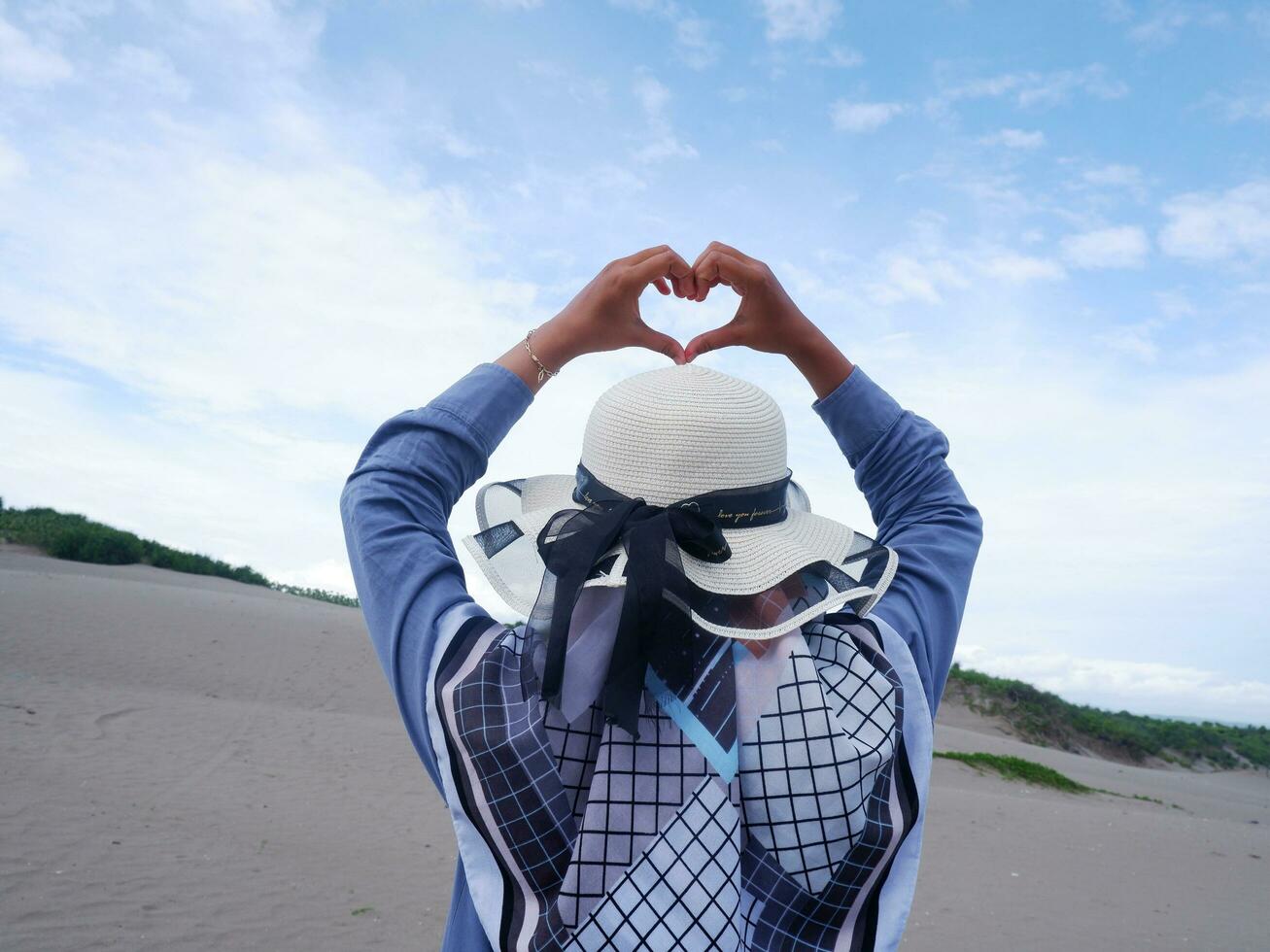 Back of the woman in the hat on the tropical beach who was staring at the ocean then formed a love from the hand. background of beach sand and sky. beach view photo