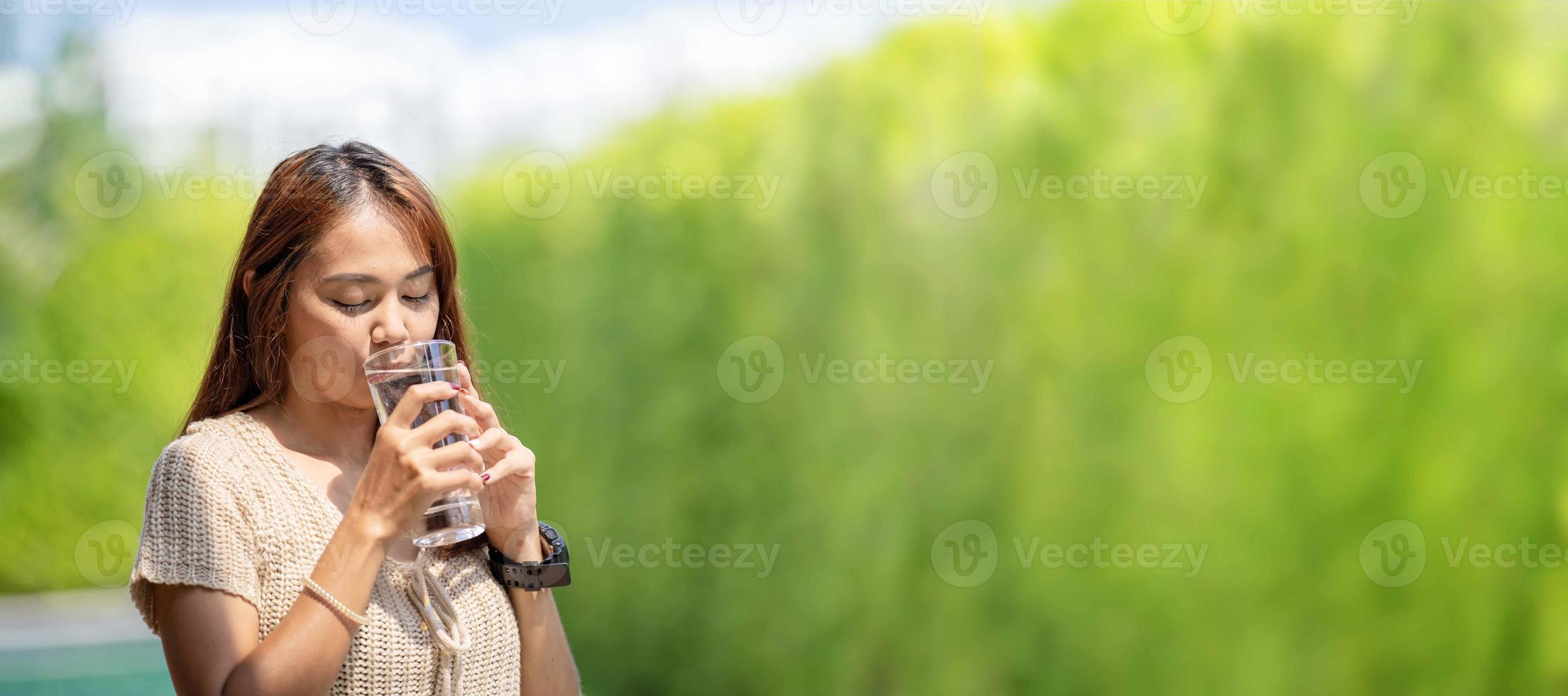 una mujer asiática bebe agua de un vaso alto de agua en el verde bokeh del árbol al aire libre para el fondo de la pancarta. foto