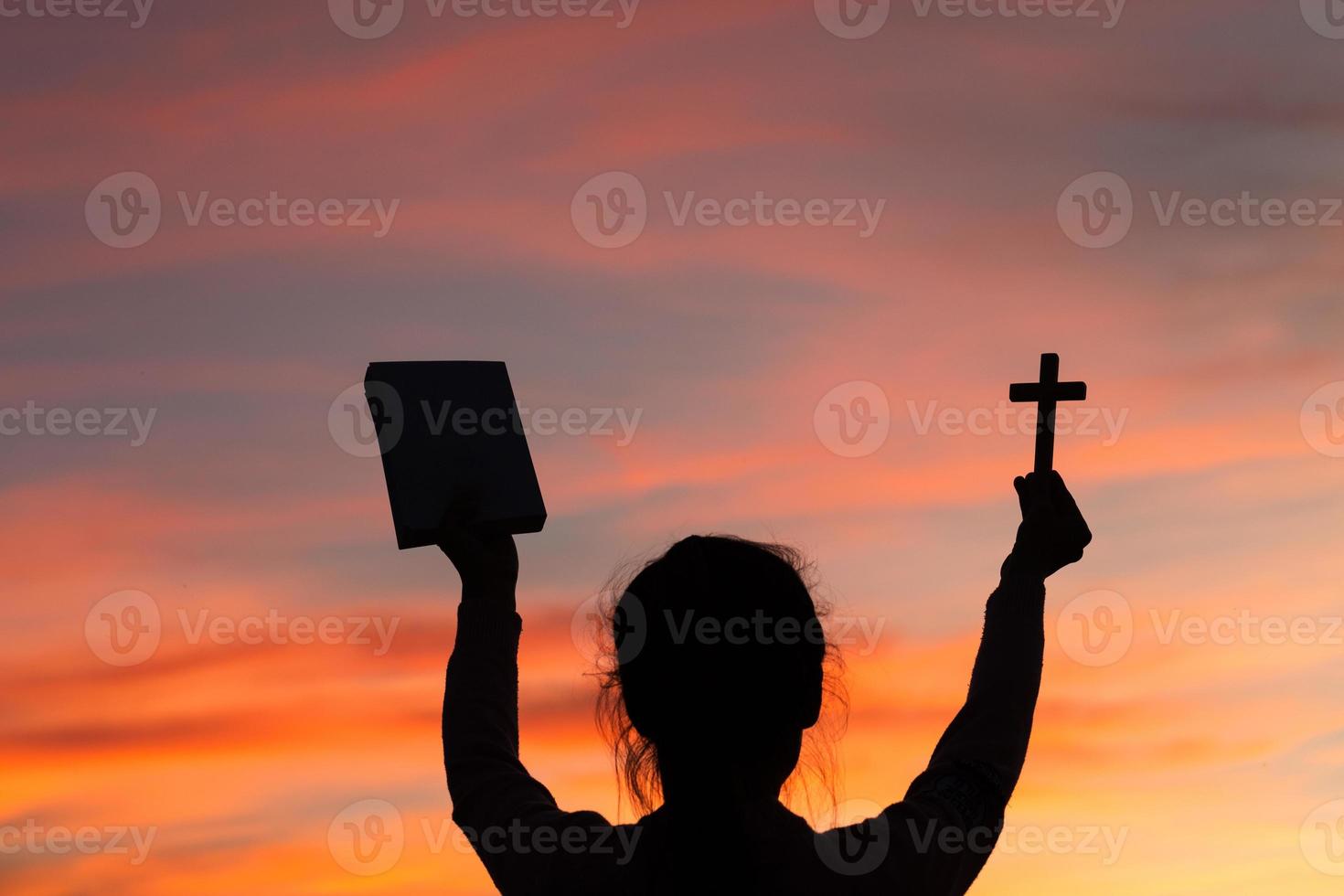 Silhouette of young woman hands holding holy Bible and lift of christian cross with light sunset background. christian concept photo