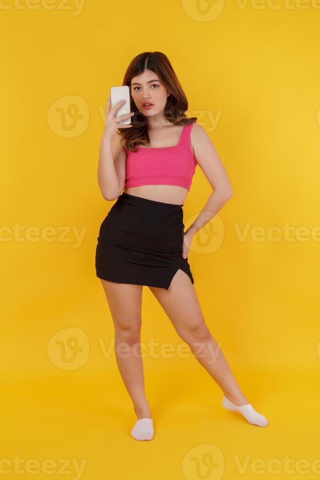 Portrait of happy young woman using mobile phone in her hands, female taking a photo with smartphone while standing isolated over yellow background