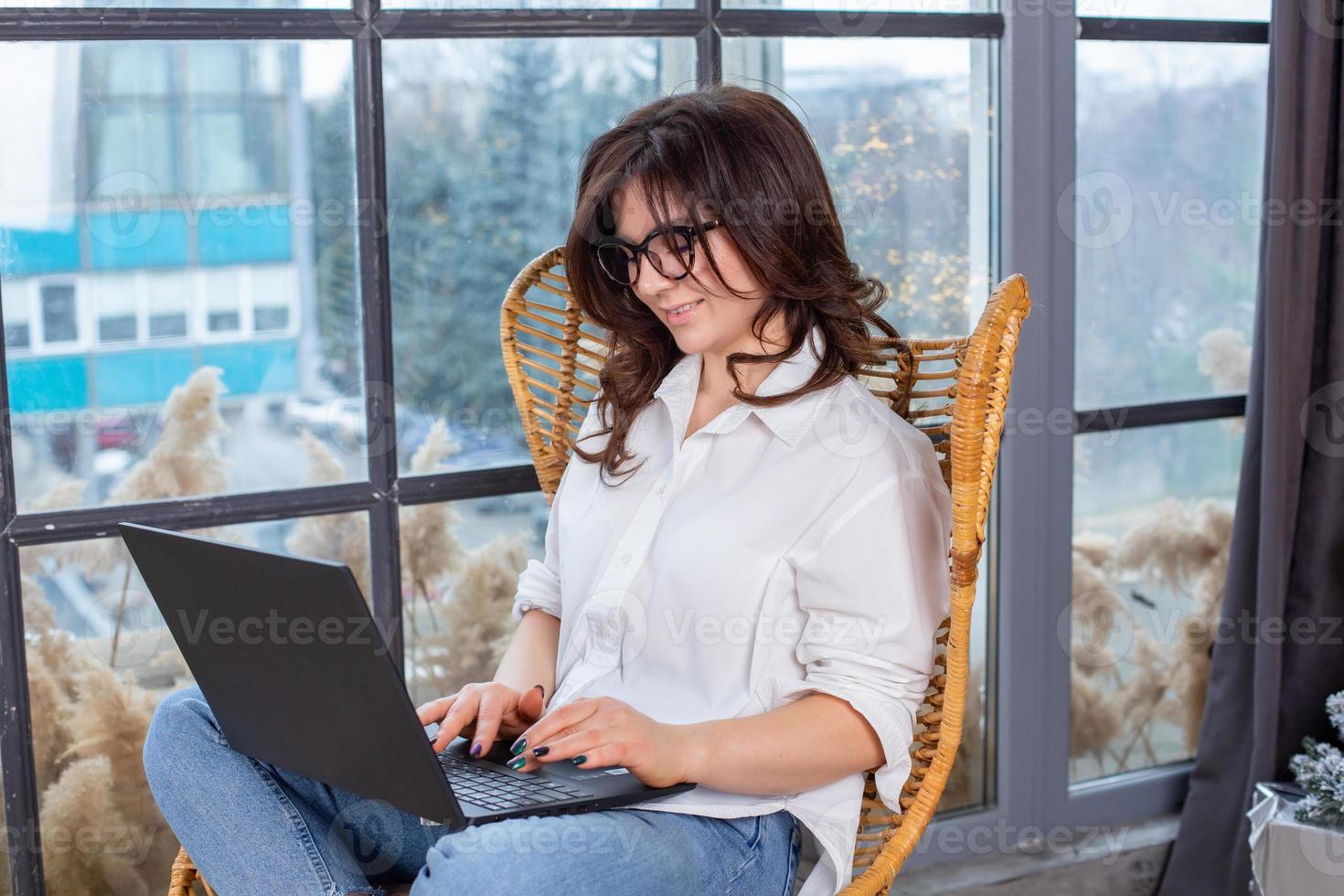 Beautiful businesswoman working on a laptop in a chair near the window. Female freelancer connecting to internet via computer. Blogger or journalist writing new article. photo