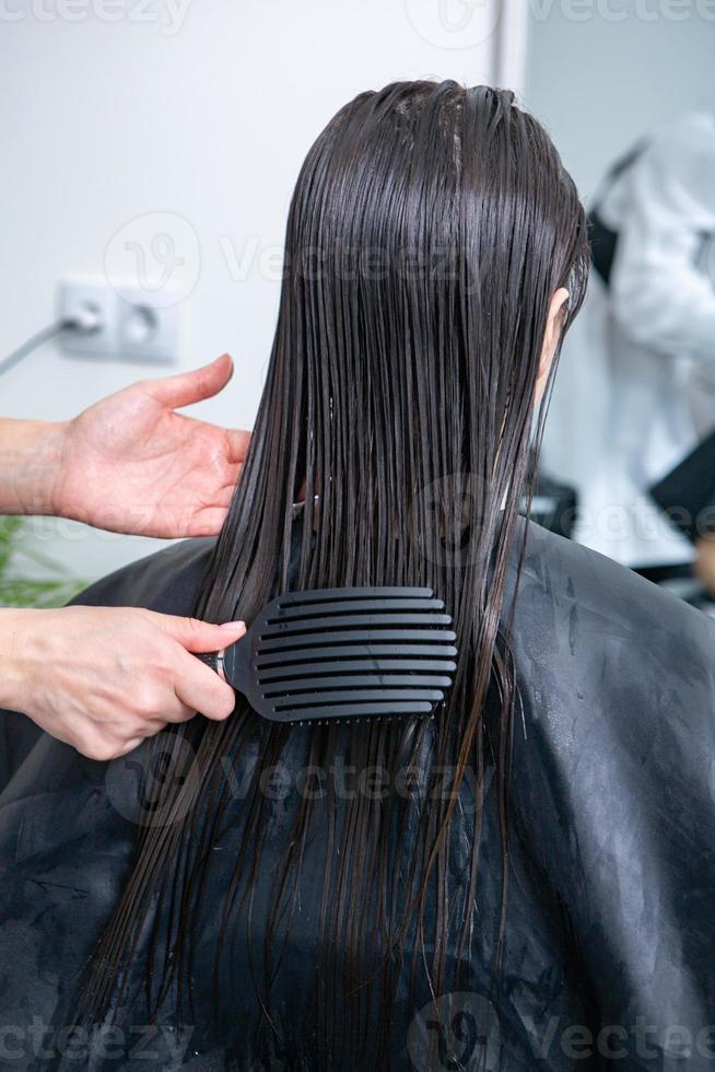 el peluquero aplica una máscara para el cabello al cabello negro liso. cuidado del cabello en el salón de belleza. foto