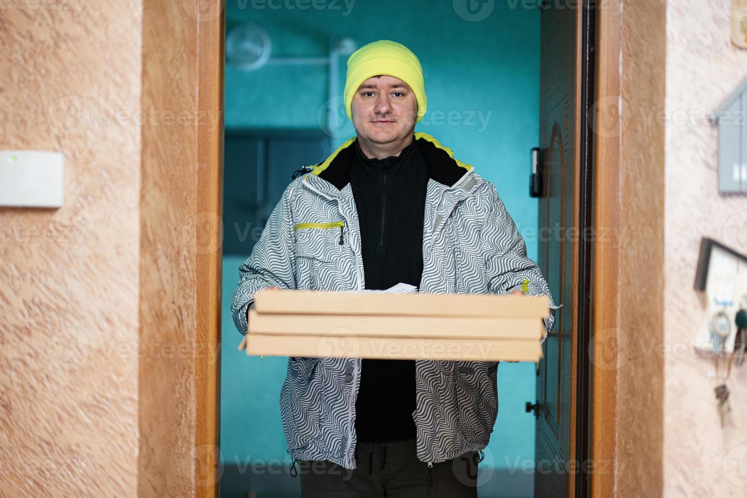 Delivery man with pizza cardboard boxes. Courier in green color hat standing against door of residential house photo