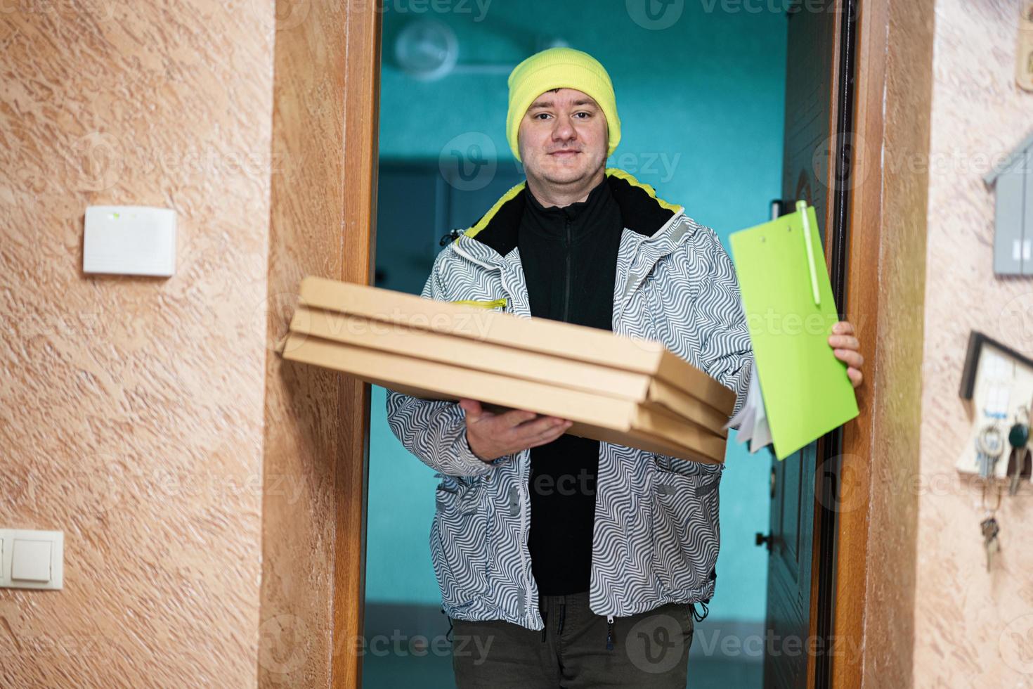 repartidor con cajas de cartón de pizza. mensajero con sombrero de color verde sosteniendo un portapapeles mientras está de pie contra la puerta de una casa residencial foto