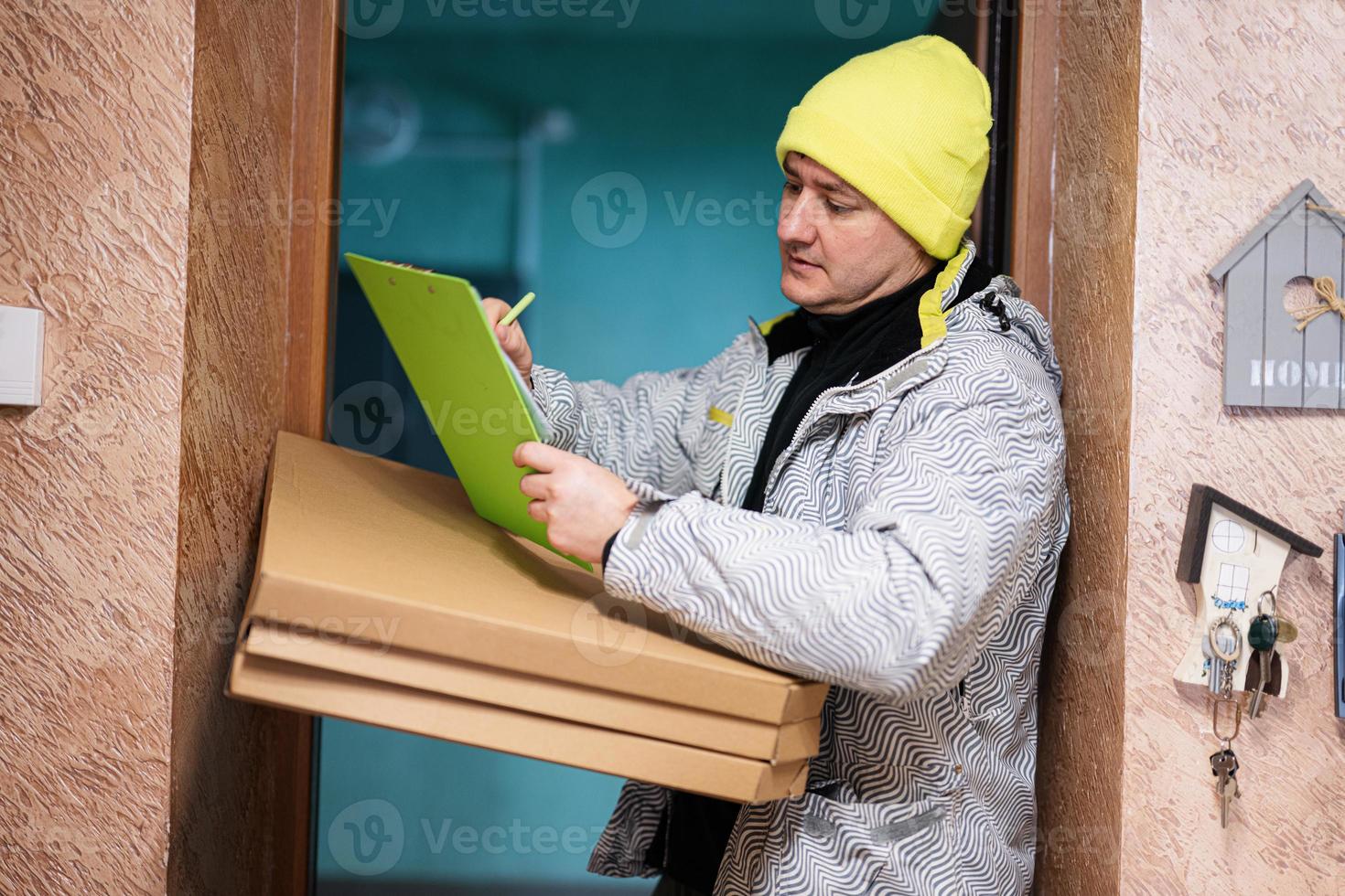 Delivery man with pizza cardboard boxes. Courier in green color hat holding a clipboard while standing against door of residential house photo