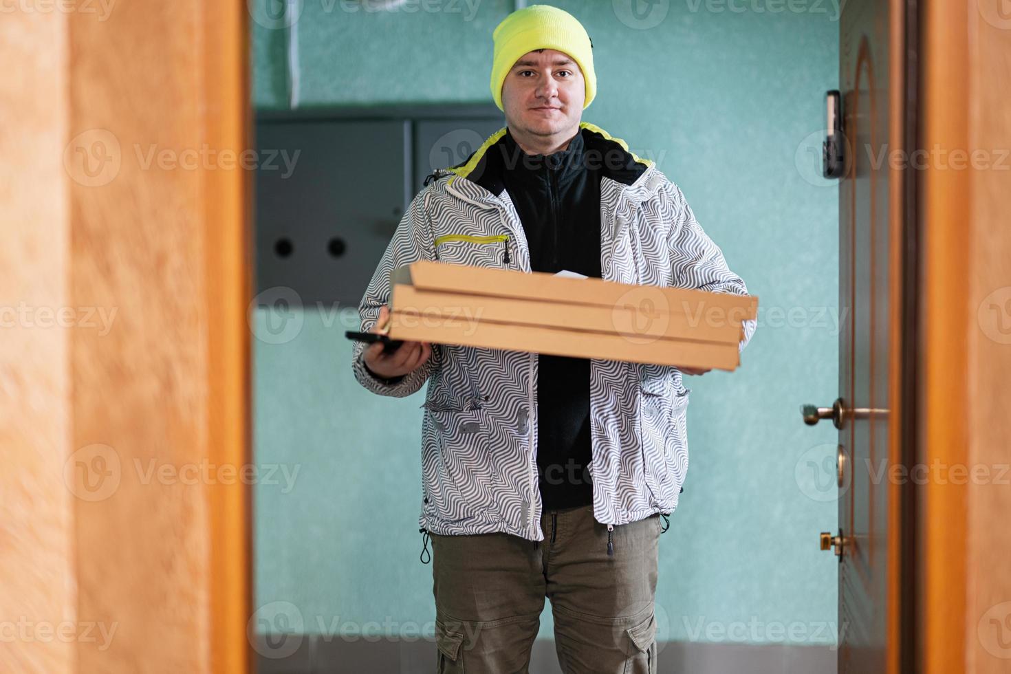 Delivery man with pizza cardboard boxes. Courier in green color hat standing against door of residential house photo