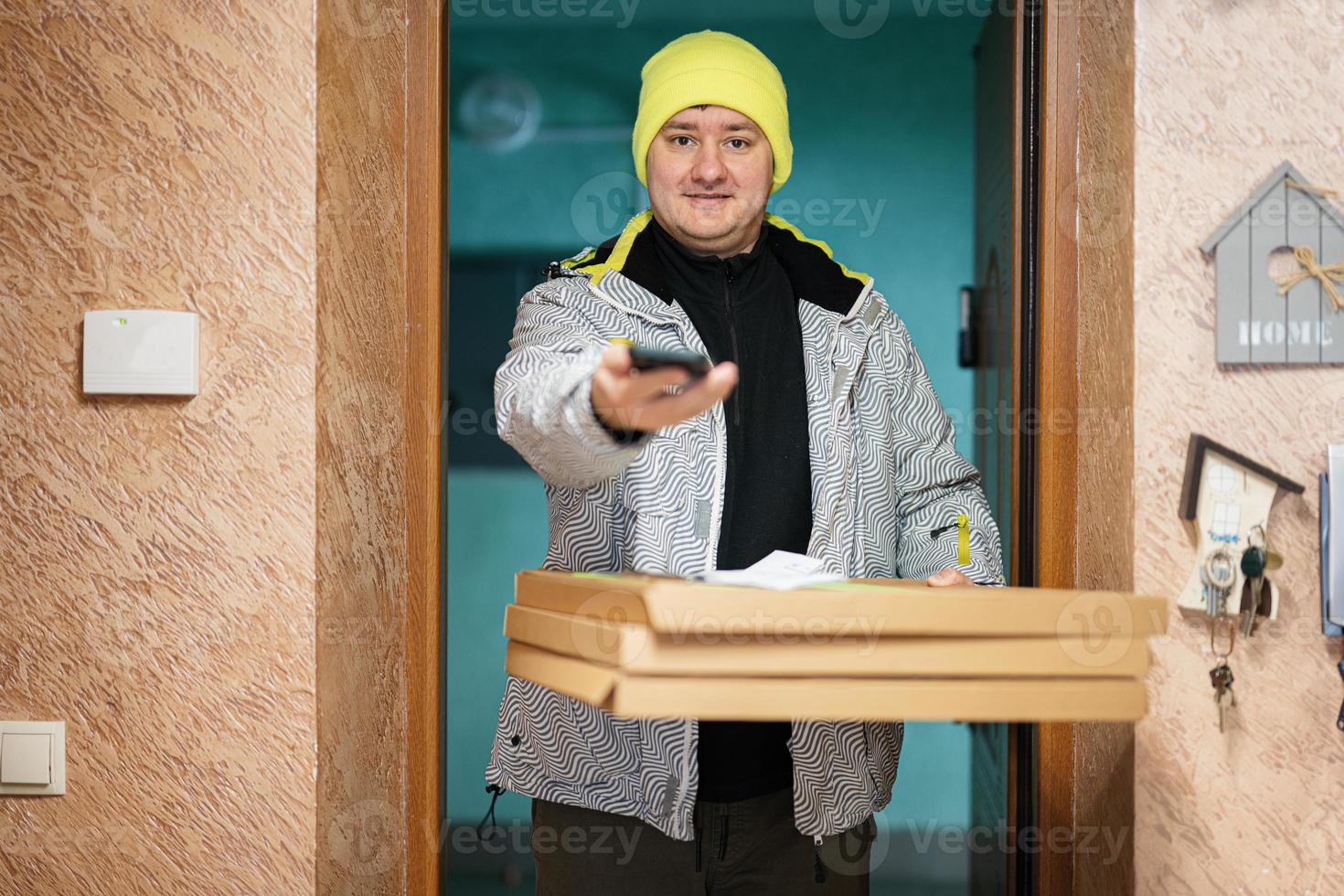 Delivery man with pizza cardboard boxes. Courier in green color hat with mobile phone standing against door of residential house photo