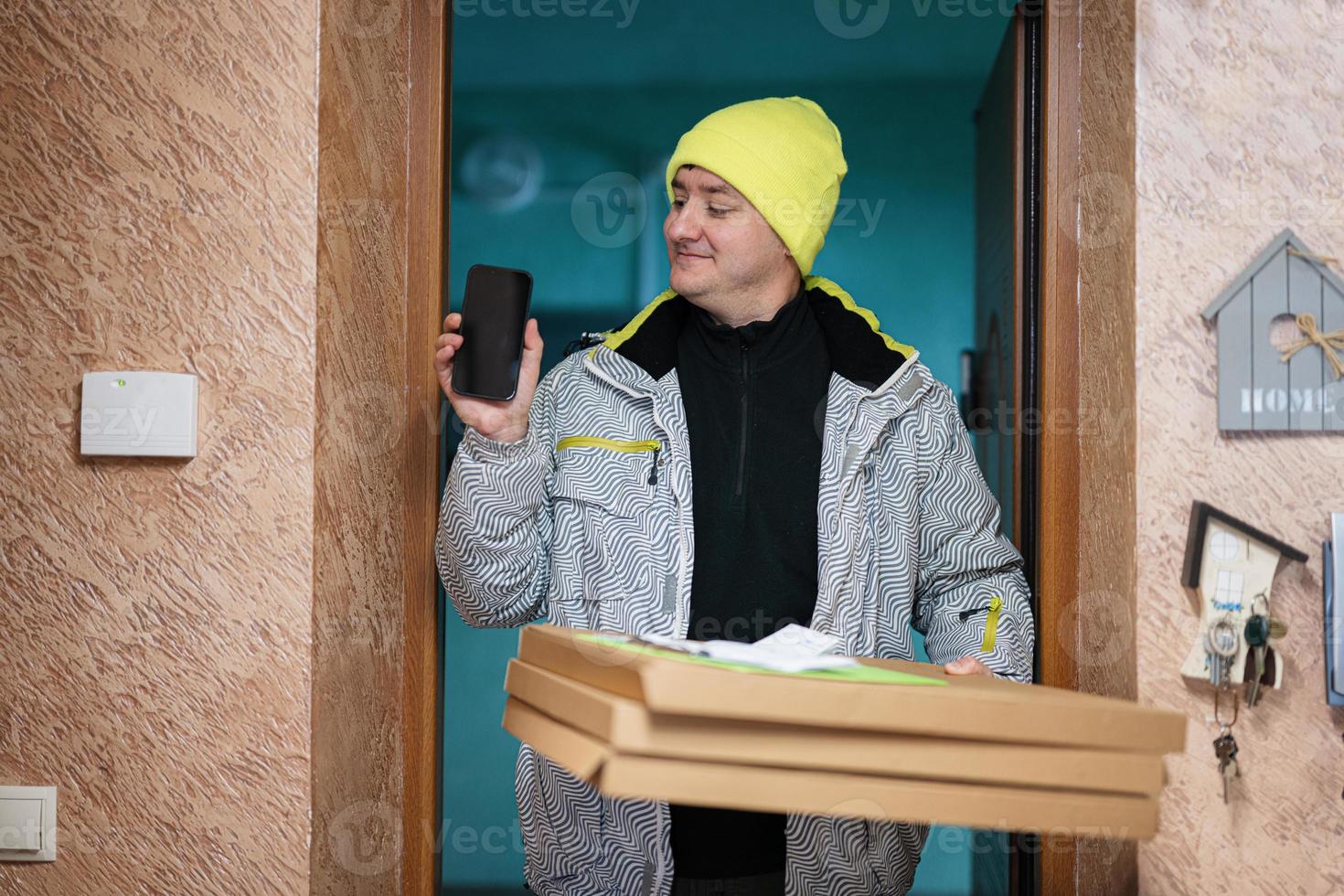 Delivery man with pizza cardboard boxes. Courier in green color hat with mobile phone standing against door of residential house photo